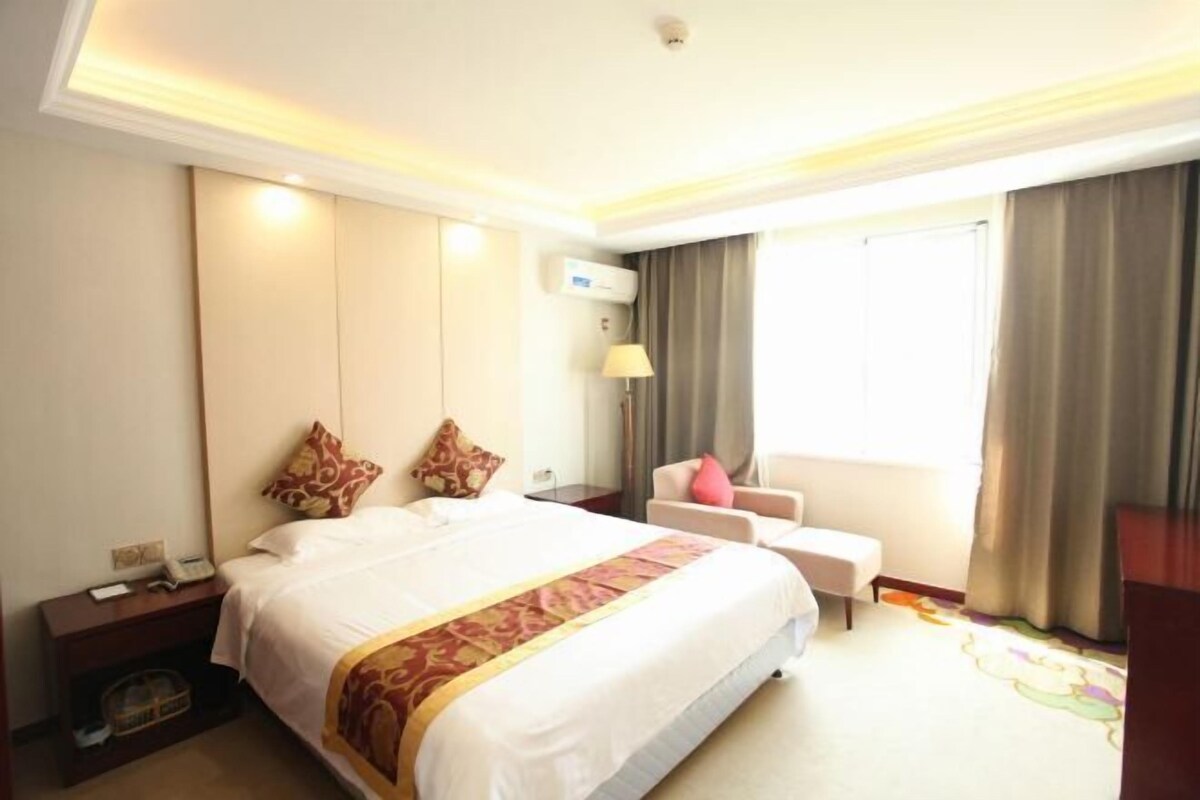 A hotel room with a large bed, a desk with a lamp, a chair, and a window with curtains.