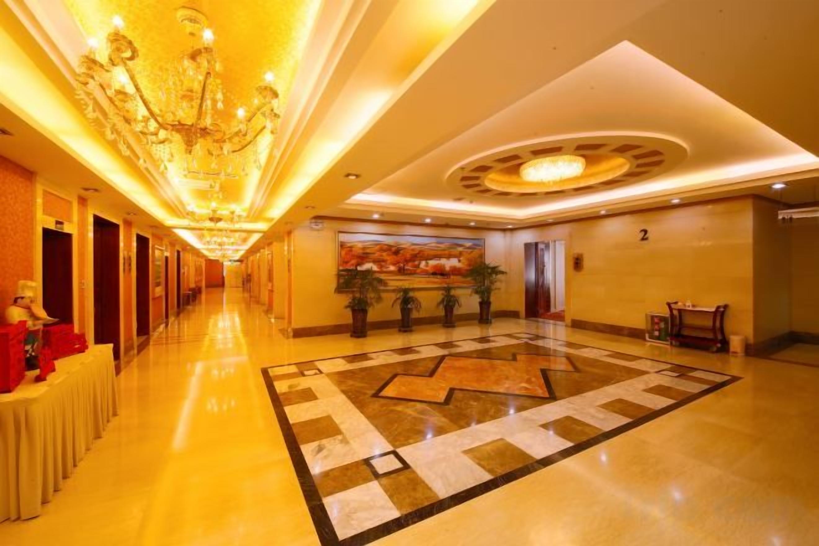 A hotel lobby with a long corridor, a chandelier, a large painting, and a patterned floor.
