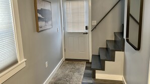 Un pasillo con una puerta blanca, una escalera de escalones oscuros, una alfombra gris y un cuadro en la pared.