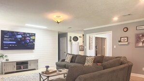 A living room with a gray sectional sofa, a flat-screen TV mounted on the wall, a coffee table, and a patterned rug. - Luxe 5BR Family Retreat, Minutes From TAMU (College Station)