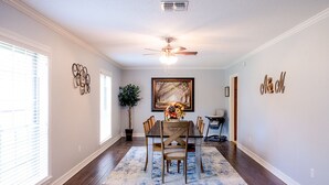 A dining area with a wooden table and chairs, a ceiling fan, a painting on the wall, and a potted plant. - Luxe 5BR Family Retreat, Minutes From TAMU (College Station)