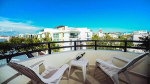 A balcony with two white lounge chairs, a small table, and a bottle of wine. The balcony overlooks a cityscape with modern buildings and a clear blue sky. - Encanto Riviera (Playa del Carmen)