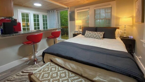 Une chambre avec un grand lit, un bureau avec deux chaises rouges, une fenêtre avec des stores et une table de chevet avec une lampe.