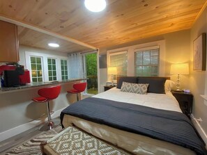 A bedroom with a large bed, a desk with two red chairs, a window with blinds, and a nightstand with a lamp.