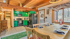 Interior de madeira com uma mesa de jantar posta para seis, armários verdes, uma geladeira e vista para uma floresta pela janela.