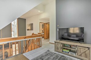 A spacious living area with a large TV, wooden entertainment unit, and a view of the outdoors through a balcony. - 4 Mi to Tubing: Cozy Bushkill Cabin w/ Deck! (Bushkill)