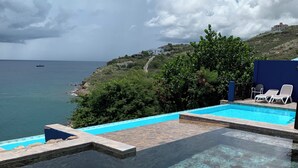 Ein Pool ohne Ende mit klarem Wasser, Blick auf das Meer und eine Hügelside mit Gebäuden.