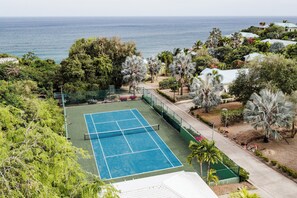A tennis court with a net, surrounded by greenery and a view of the ocean.