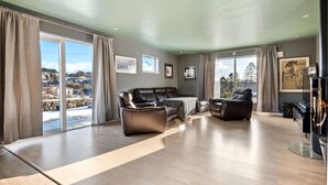 A spacious living room with a large sofa, a recliner, and a coffee table. There are large windows with curtains, a fireplace, and framed artwork on the walls.