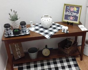 Una mesa de madera con un mantel a cuadros blanco y negro, una jarra de cerámica blanca, una pequeña planta en maceta y un cartel que dice 'welcome'.