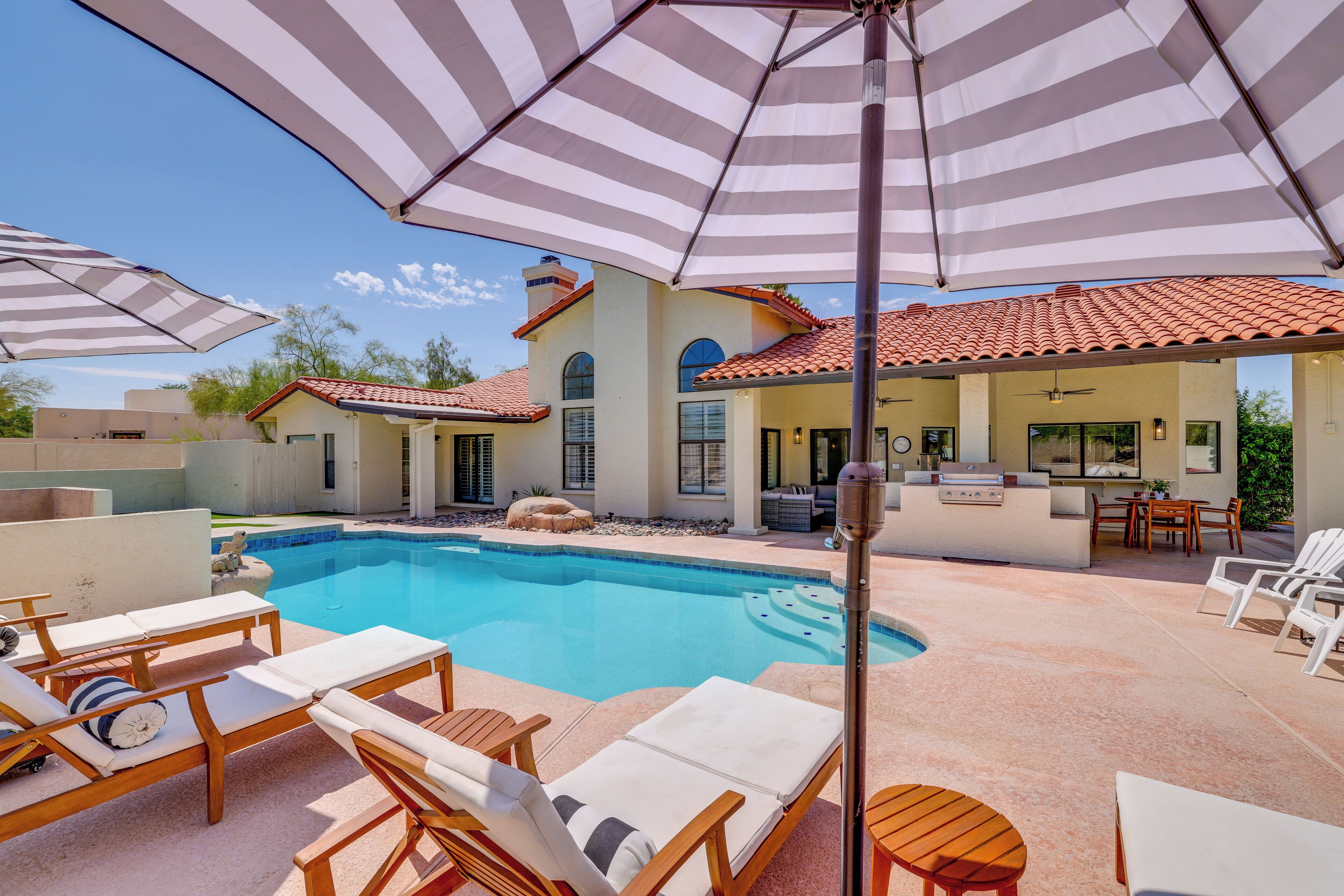 A poolside area with lounge chairs, a barbecue station, and umbrellas providing shade.