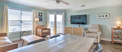 A spacious living room with a large wooden dining table, two sofas, a ceiling fan, and a television mounted on the wall.
