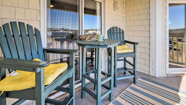 A covered porch with two Adirondack chairs and a small table, overlooking a scenic view. - Oak Island Condo - Walk to the Beach! (Oak Island)