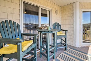 A covered porch with two Adirondack chairs and a small table, overlooking a scenic view. - Oak Island Condo - Walk to the Beach! (Oak Island)
