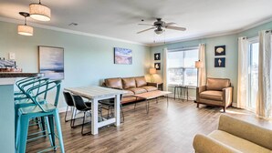 A living room with a sofa, chairs, a coffee table, a dining table with chairs, a ceiling fan, and a window with curtains. - Oak Island Condo - Walk to the Beach! (Oak Island)