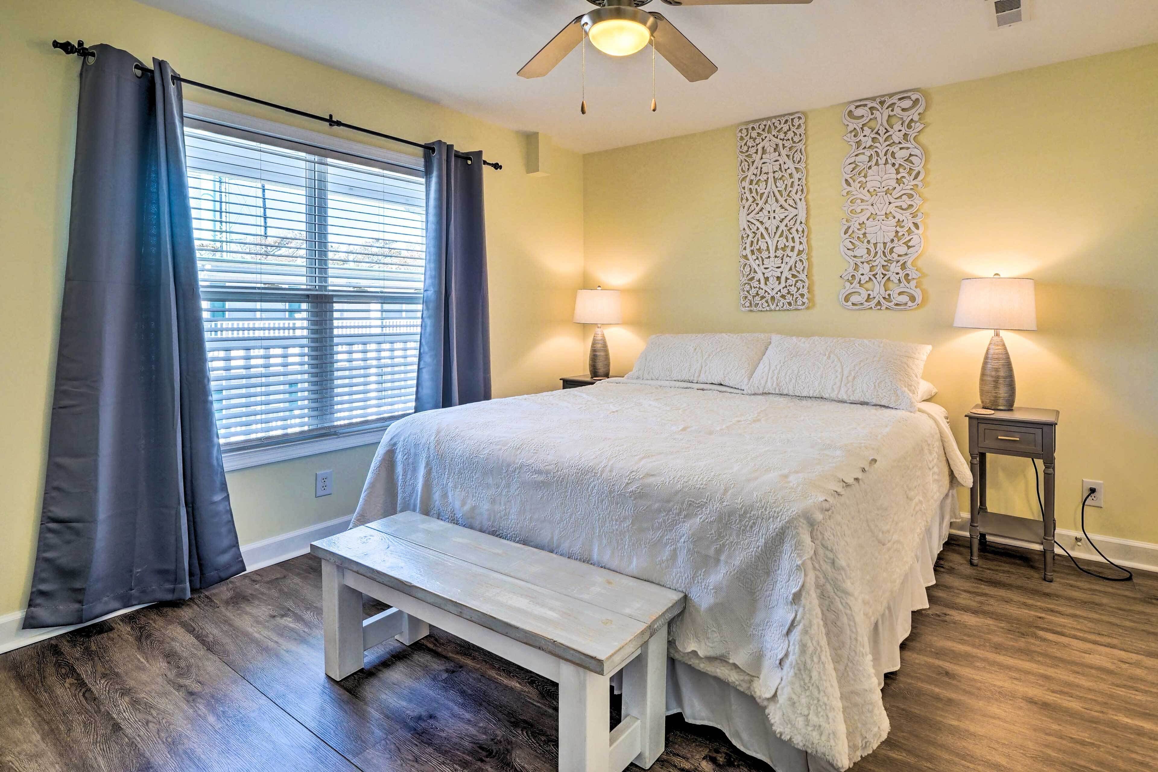 A bedroom with a bed, a bench, two bedside lamps, a ceiling fan, and decorative wall art.