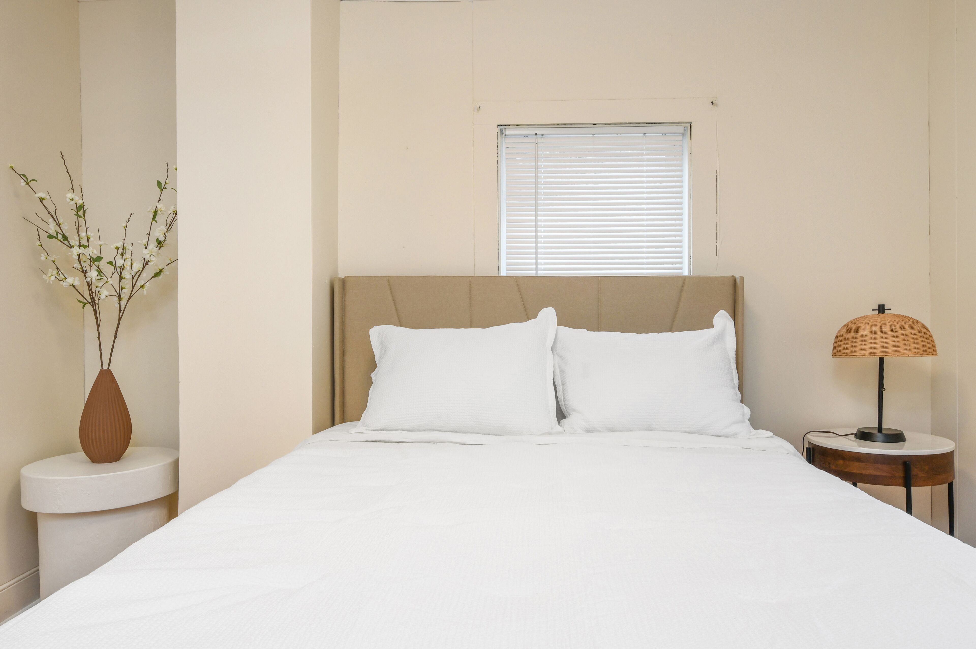 A bedroom with a bed, white bedding, a window with blinds, a bedside table with a lamp, and a vase with branches.