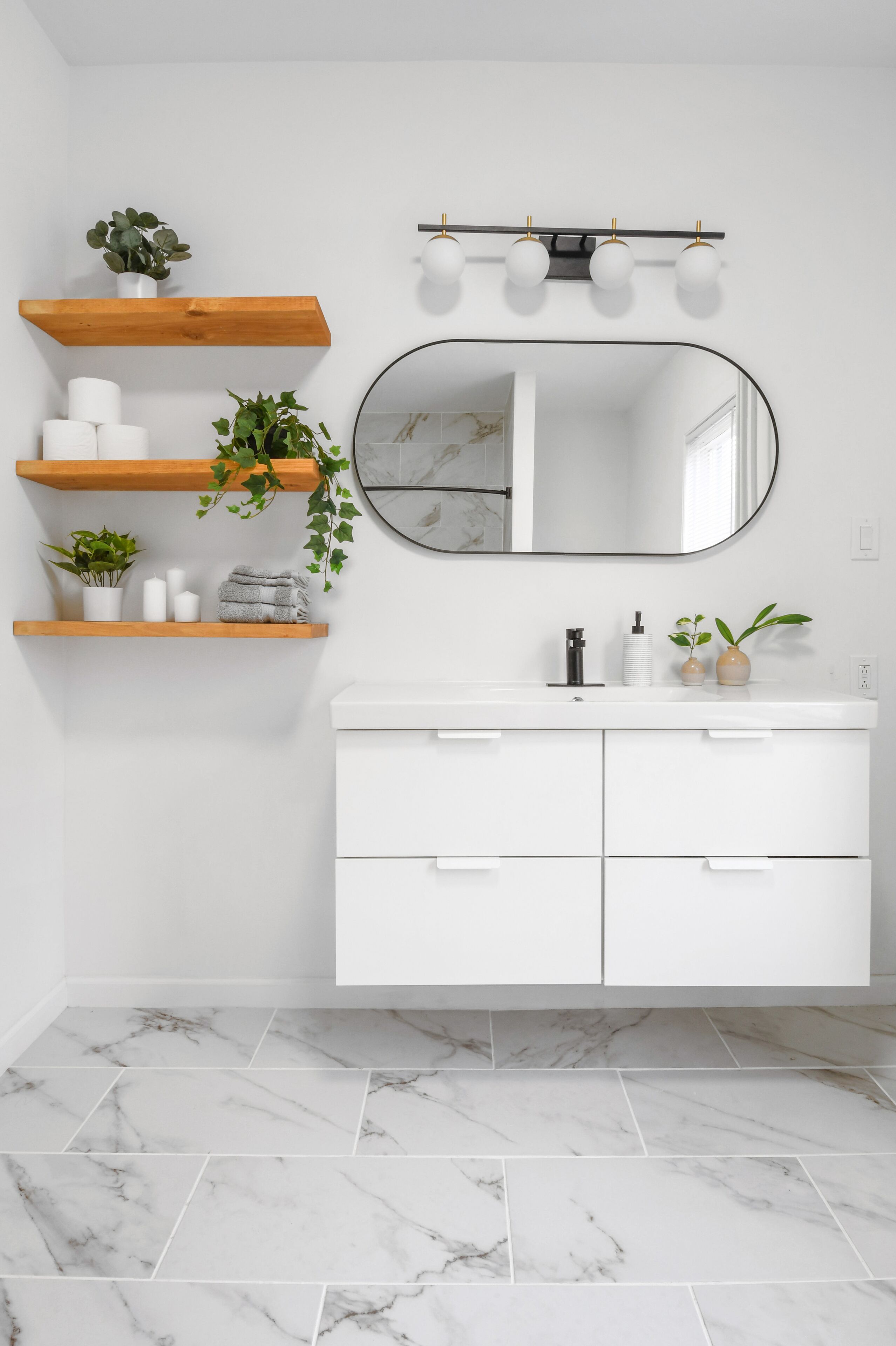 Une salle de bain avec un meuble-lavabo blanc, des carreaux de marbre, des étagères en bois ornées de plantes et un miroir avec éclairage intégré.