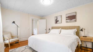 Un dormitorio con una cama grande, una silla, una mesita de noche con lámpara y dos cuadros colgados en la pared.