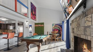 A pool table in a spacious room with a fireplace, a kitchen area, and a bar.