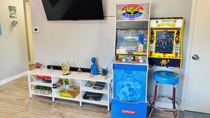 Una habitación con una televisión de pantalla plana montada en la pared, un estante con juegos de mesa, una máquina arcade Capcom y un taburete azul con temática Capcom.
