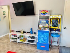 Una habitación con una televisión de pantalla plana montada en la pared, un estante con juegos de mesa, una máquina arcade Capcom y un taburete azul con temática Capcom.