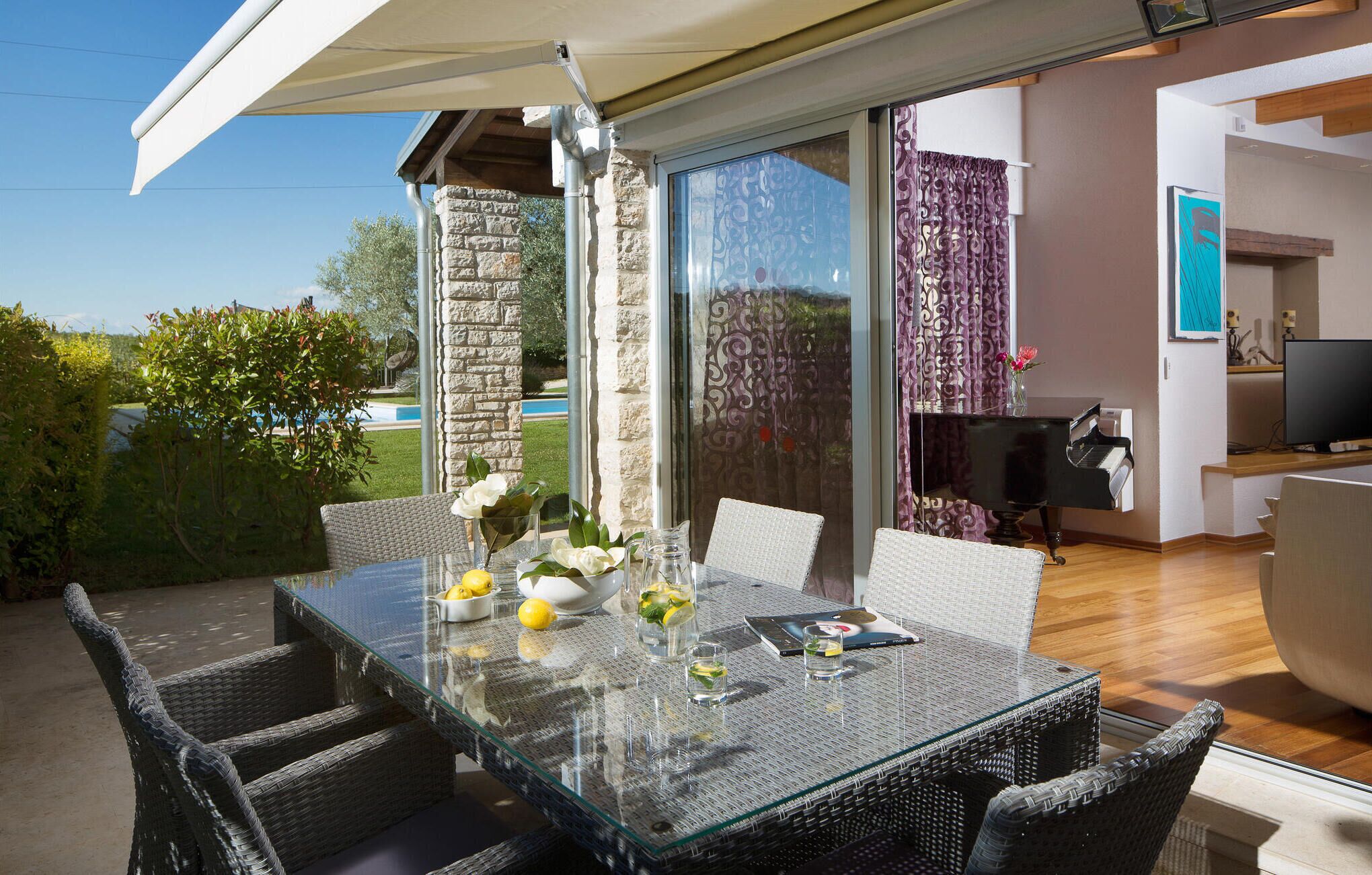 Ein Esstisch mit Glasplatte, Korbstühle, ein Klavier und ein Fernseher in einer modernen Wohnarea mit Blick auf einen Pool.