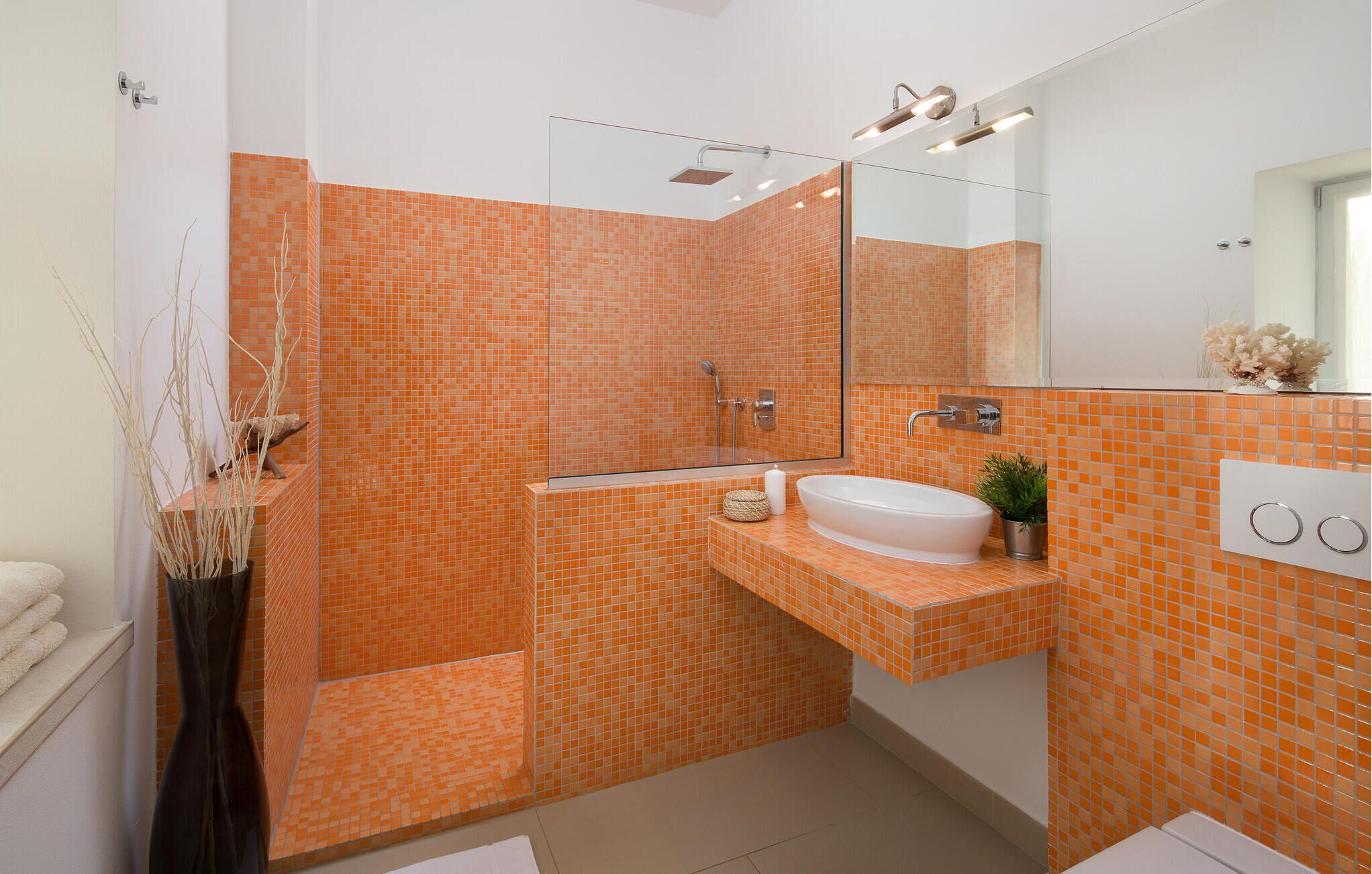 Un baño con mosaicos anaranjados, ducha con cerramiento de vidrio, lavamanos blanco y un espejo.