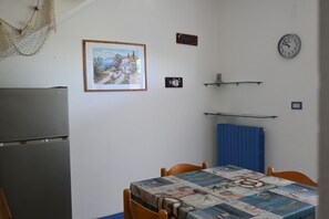 A small kitchen with a refrigerator, a dining table with chairs, a clock, and a framed picture on the wall.