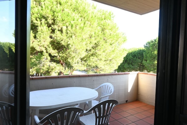 An outdoor patio with a white table and chairs, a tiled floor, and a view of a large tree and shrubbery.