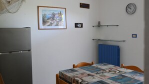 A small kitchen with a refrigerator, a dining table with chairs, a clock, and a framed picture on the wall. - M328 - Marcelli, two-room apartment with sea view (Marcelli)