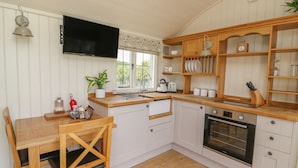 A compact kitchen with white cabinetry, a wooden dining table, and a built-in oven. - Hillside View Shepherds Hut Sleeps 2 (Rhayader)