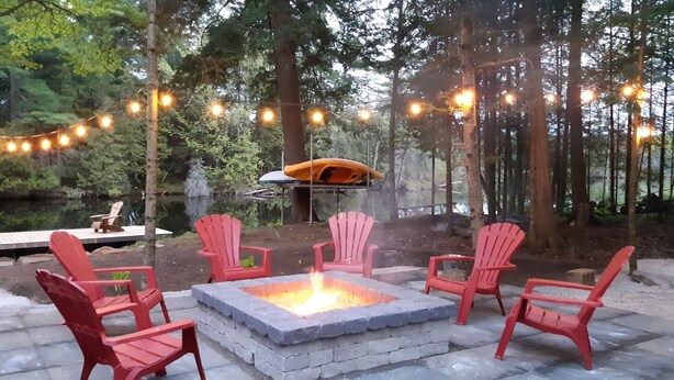 A fire pit with red chairs, a covered seating area, and string lights in a forested outdoor setting. - Cozy cottage with amazing view on the Big Bald Lake in Bobcaygeon (Bobcaygeon)