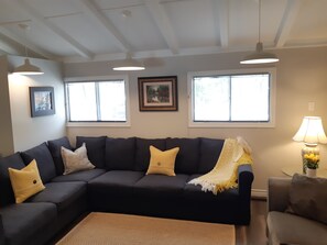 A living room with a dark-colored sectional sofa, two windows with blinds, a framed picture on the wall, and a lamp on a side table.
