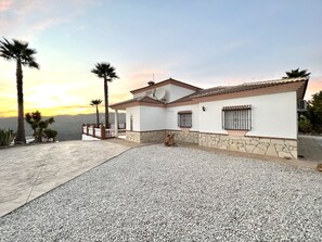 A two-story house with a stone foundation and white walls, featuring a patio area with gravel and a white fence.