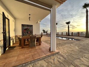 A covered patio with a dining table and chairs, a small pool, and a view of palm trees and mountains.