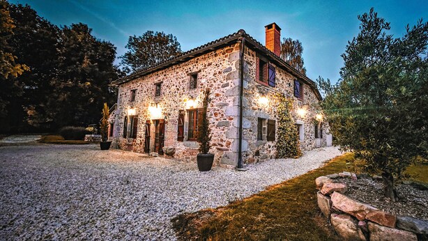 A stone building with multiple windows and a chimney, surrounded by a gravel driveway and a small tree.