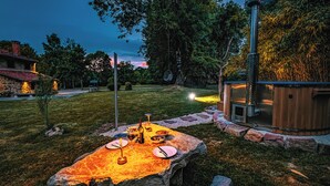 An outdoor dining area with a stone table set for a meal, surrounded by a stone wall and a wooden hot tub.
