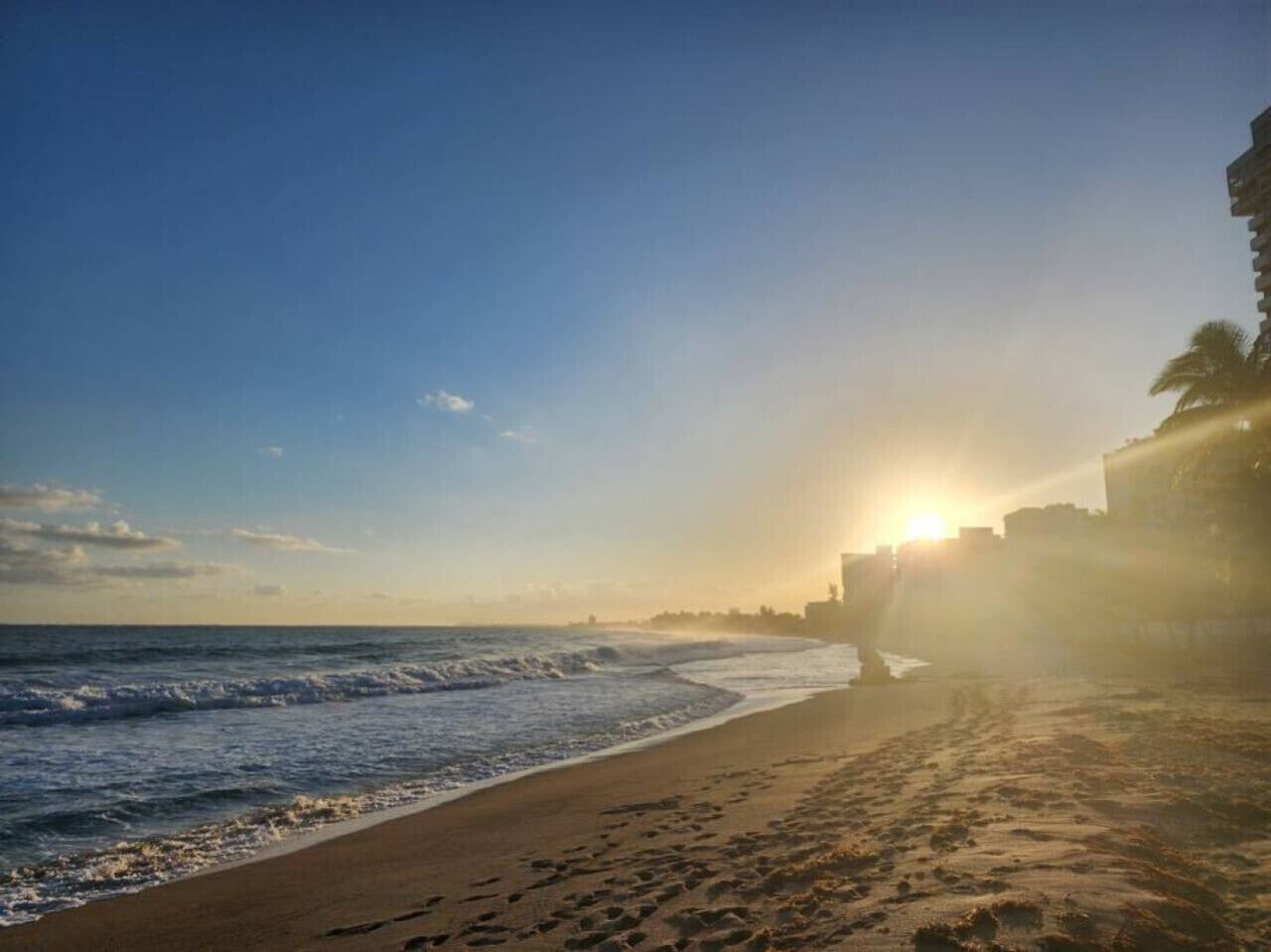 A beach with a sunset, waves, and a distant cityscape.