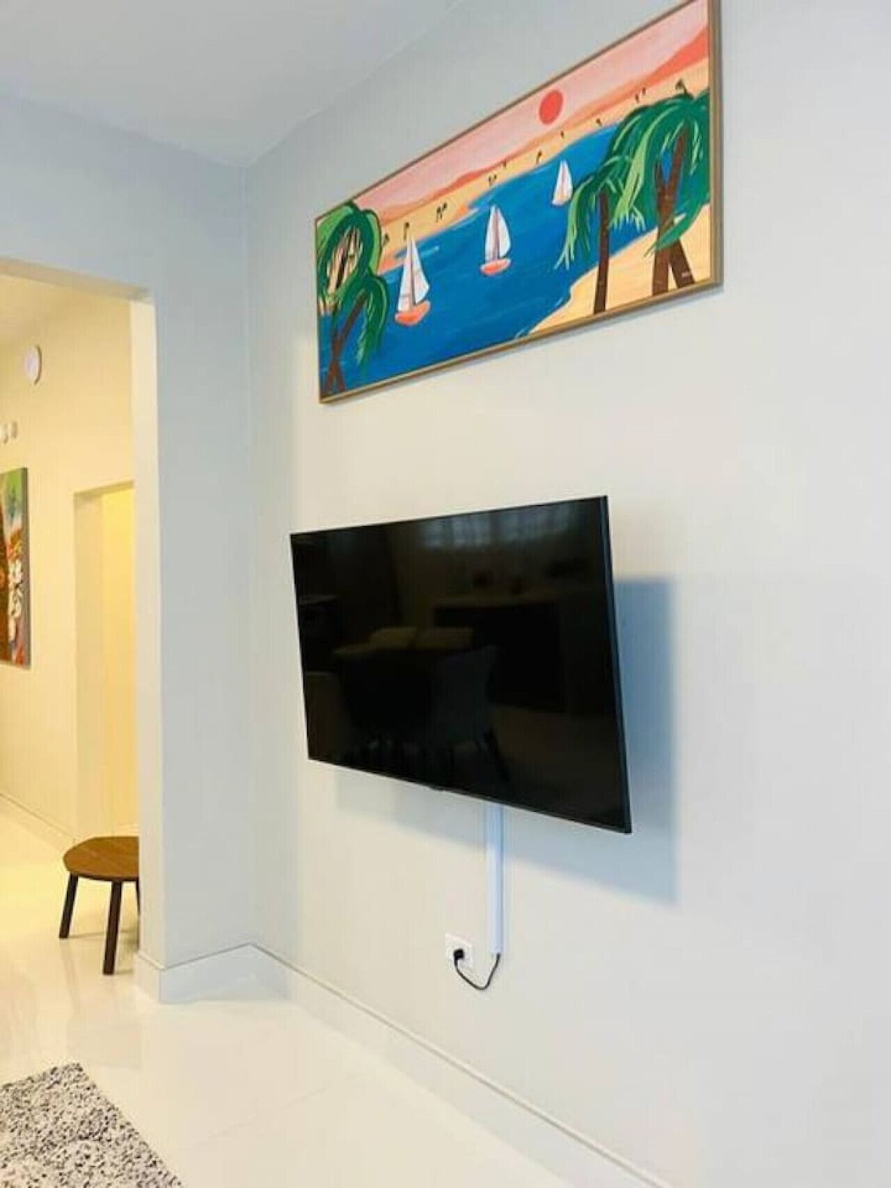 A modern living room with a flat-screen TV mounted on the wall, a painting of a sunset over water, and a small table with chairs.