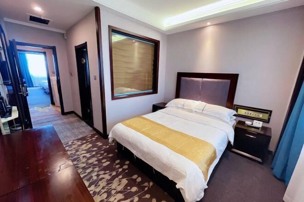 Ein Hotelzimmer mit einem großen Bett, einem Nachttisch, einem Fenster mit Jalousien und einer Tür zu einem weiteren Raum.