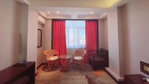 Une chambre d’hôtel dotée d  ’un coin salon avec des fauteuils en osier et une petite table ronde, un canapé sombre et une grande fenêtre ornée de rideaux rouges.