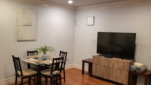 Een eethoek met een houten tafel, stoelen en een potplant. Een flatscreen televisie is bevestigd aan een houten kast, en een klein tafeltje met een afstandsbediening staat ernaast.