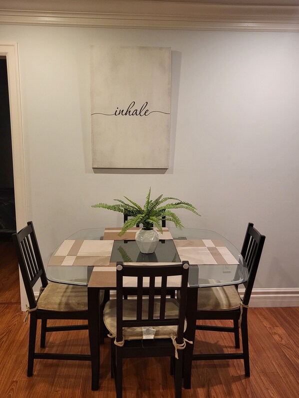 A dining table with a glass top, surrounded by four wooden chairs. A potted plant is on the table, and a wall-mounted sign above reads "inhale".