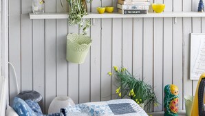 Una habitación con pared de madera blanca, cama con colcha azul y blanca, planta colgante verde, planta pequeña en maceta y estante con libros y objetos decorativos.