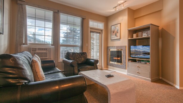 A living room with a fireplace, leather sofa, and a television on a stand. - Mountain Rook 2 king bedroom, sleeps 6: Unit 1002 (Fairmont Hot Springs)