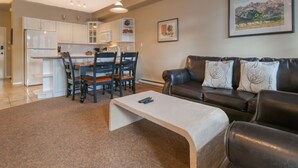 A modern living room with a brown leather sofa, a coffee table, and a dining area with a kitchen in the background. - Mountain Rook 2 king bedroom, sleeps 6: Unit 1002 (Fairmont Hot Springs)