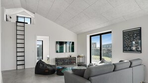 A modern living room with a grey sofa, a black bean bag, a glass coffee table, and a TV on the wall. There is a ladder in the corner and a large window with a view of the outside.