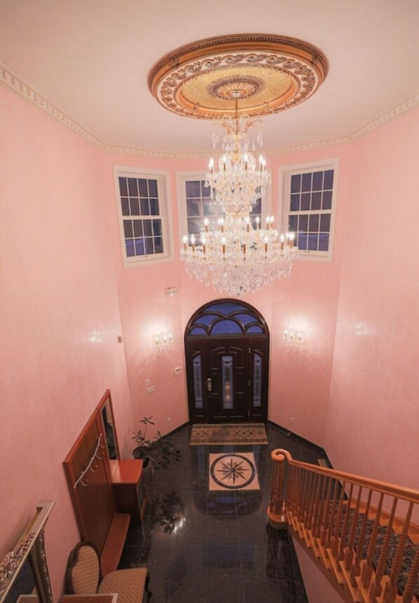 A grand staircase with a chandelier, a wooden handrail, and a black tiled floor leading to a dark wooden door with a decorative arch.