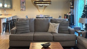 A living room with a sofa, coffee table, and a ceiling fan. There is a kitchen area with bar stools and a sign that reads "Welcome".